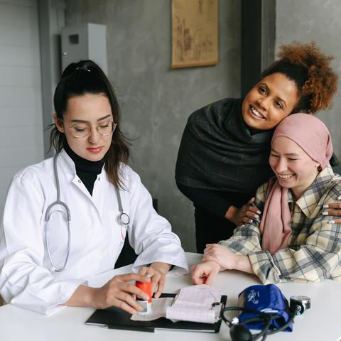 Eine Ärztin sitzt am Tisch in einem Beratungsgespräch mit zwei Patientinnen. Auf dem Tisch liegt ein Blutdruckmessgerät. Alle Frauen lächeln. 
