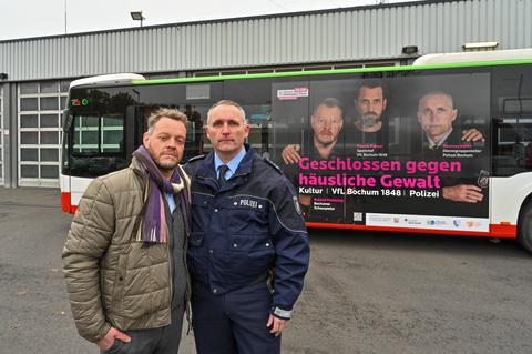 Schauspieler Roland Riebeling und Polizist Andreas Derks stehen  vor einem Bus mit dem Motiv einer Kampagne gegen häusliche Gewalt, für das sie gemeinsam mit VfL-Sportchef Patrick Fabian fotografiert wurden. 