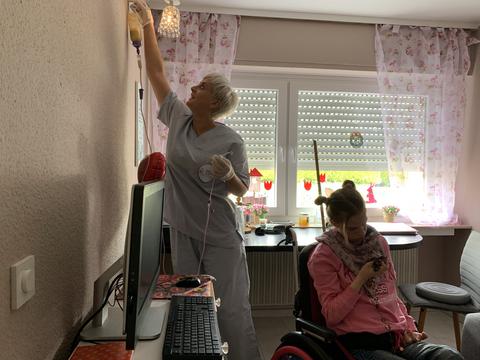 Eine Pflegekraft platziert an einer Wandhalterung eine Ernährungsflasche für eine im Rollstuhl sitzende junge Frau.