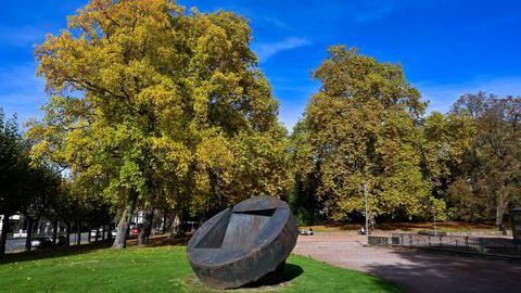 Große Metallskulptur auf einer Wiese mit herbstlichen Bäumen.